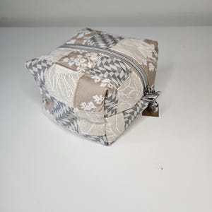 Könnte beinhalten: Eine quadratische, gesteppte Kosmetiktasche mit Patchwork-Design. Die Tasche hat einen silbernen Reißverschluss und eine Mischung aus beigen, grauen und weißen Stoffquadraten mit floralen und geometrischen Mustern. Die Tasche ist so konzipiert, dass sie aufrecht steht.