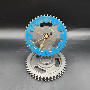 May include: A table clock constructed from two stacked metal gears. The top gear features a blue circular ring with the numbers 1 through 12 printed in blue. Gold-colored clock hands are centered on the gear, which is marked with the text S468.