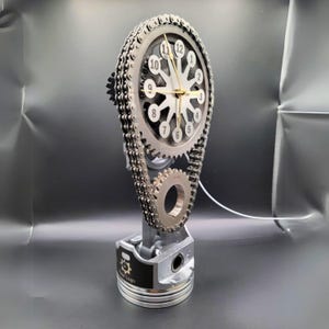May include: A silver and black clock with a chain and gear design. The clock face has a white background with black numbers and a gold hand. The clock is mounted on a silver piston.