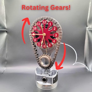 May include: A red and silver clock with a rotating gear design. The clock face is made of metal and has a red background with white numbers. The clock is mounted on a silver piston with a chain and gear mechanism. The clock is titled "Rotating Gears!"