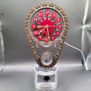May include: A clock with a red face and black numbers is mounted on a silver metal piston and chain. The clock is a unique and stylish way to add a touch of industrial chic to any room.