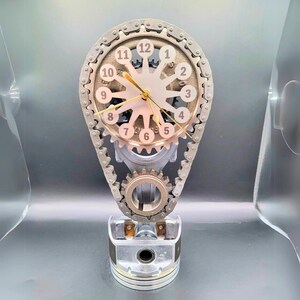 May include: A silver and gold clock with a chain and gears, resembling a car engine, with a white face and black numbers. The clock is sitting on a silver piston.