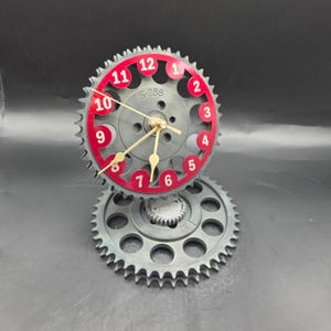 May include: A unique clock made from repurposed gears. The clock face is a gray gear with red numbers and a red outer ring. The clock hands are gold. The clock sits on top of a larger gear, creating a distinctive industrial design.