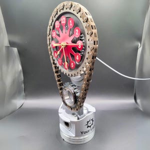 May include: A unique clock made from repurposed engine parts. The clock face is red with white numbers and gold hands, surrounded by a metal chain. The base is a silver piston with the text "Your Logo".
