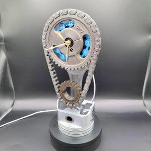 May include: A clock made from a car engine piston, connecting rod, and timing chain. The clock face is a silver metal disc with blue numbers and a gold-colored hour and minute hand. The clock is mounted on a black base.