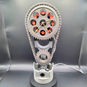 May include: A clock made from a car engine crankshaft, connecting rod, and piston. The clock face is a silver gear with orange numbers and hands. The clock is mounted on a black base.