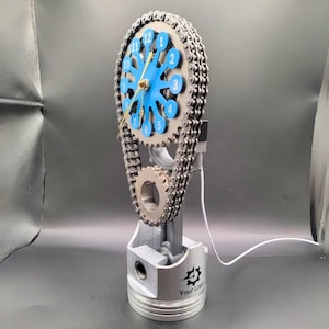 May include: A clock made from a car engine piston, chain, and gears. The clock face is blue with white numbers and a gold hand. The base of the clock is a silver piston with the text "Your Logo" on it.