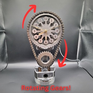 May include: A clock made from a car engine part with a silver face and black numbers. The clock is mounted on a piston and has a chain drive that rotates the gears. The clock face reads "Rotating Gears!"
