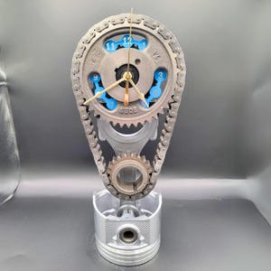 May include: A clock made from a car engine crankshaft, connecting rod, and piston. The clock face is a silver gear with blue numbers and a gold-colored hour and minute hand. The clock is mounted on a silver piston.