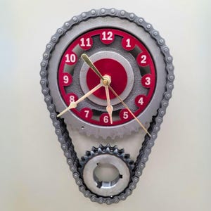 May include: A clock made from metal gears and a chain. The clock face is red and silver with black numbers. The clock hands are gold.