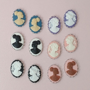 May include: A collection of oval cameo pendants in various colors, including blue, purple, pink, and black. Each cameo features a profile of a woman with detailed hairstyles, set within a silver-toned frame. The cameos are arranged on a light pink surface.