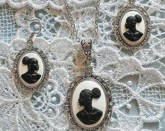 May include: A jewelry set consisting of a necklace and two matching earrings, each featuring an oval cameo pendant with a black silhouette of a person with an afro hairstyle set against a cream background, framed in ornate silver-toned metal.