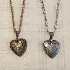 Peut inclure: Deux colliers avec pendentifs en forme de cœur. L'un a une chaîne et un pendentif dorés avec un motif orné. L'autre a une chaîne et un pendentif argentés avec un motif similaire. Les deux pendentifs ont un cœur texturé au centre.