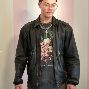 May include: A young person wearing a black leather jacket with a zipper closure. They are wearing a grey t-shirt with a skull graphic and the text "Creative Control". They are also wearing a silver chain necklace with a pendant.