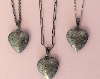 May include: Three silver-toned heart-shaped lockets on chains. Each locket has a textured center and ornate border. The chains vary in style, with one featuring a link chain. The background is a solid light pink.