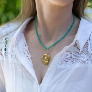 May include: A gold pendant necklace featuring a spiral design with a pearl center, suspended from a turquoise beaded chain. The necklace is worn over a white shirt with lace detailing.