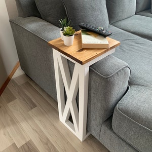 May include: A white side table with a wooden top, featuring an X-shaped design on the legs. The table is next to a gray couch with a book and a remote control on top.