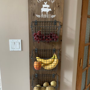 May include: A wooden wall organizer with the text "Farm Fresh Produce" and a silhouette of a chicken, pig, and cow. Three wire baskets are attached to the organizer, holding grapes, bananas, and potatoes.