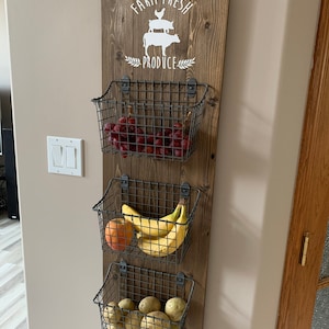 May include: A wooden wall-mounted organizer with three wire baskets. The top basket holds red grapes, the middle basket holds bananas and an apple, and the bottom basket holds potatoes. The wood has a sign that says "Farm Fresh Produce" with a silhouette of a chicken, pig, and cow.