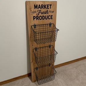 May include: A wooden wall organizer with three wire baskets. The organizer has a sign that says "MARKET Fresh PRODUCE". The wood is stained a dark walnut color. The sign is black.