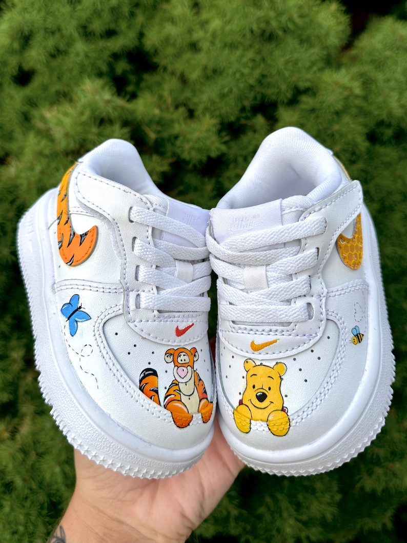 May include: White baby trainers with custom painted Winnie the Pooh characters. The shoes feature a blue butterfly, a bee, and the characters Tigger and Winnie the Pooh. The Nike swoosh is orange and yellow.