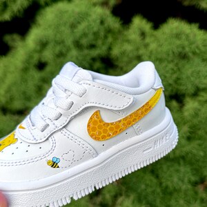 May include: White toddler trainer with a yellow and black bee and honeycomb design. The Nike swoosh is yellow with a honeycomb pattern. A cartoon bee and heart trail are on the side.