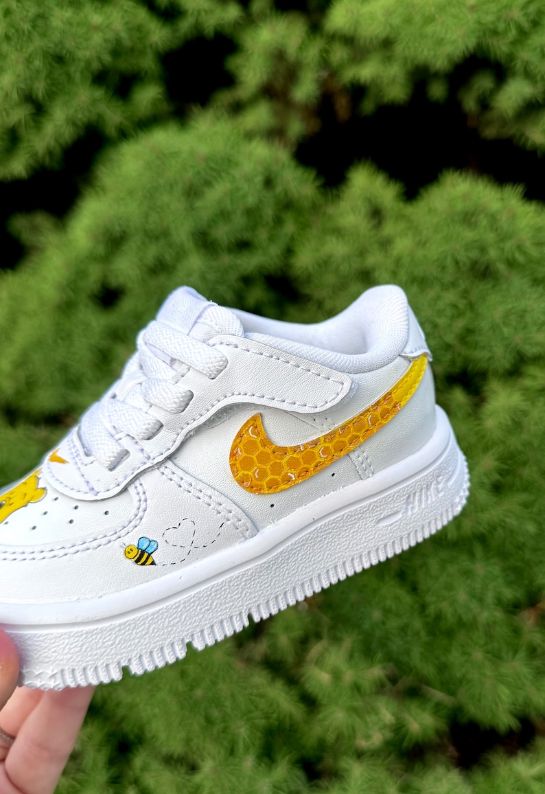 May include: White toddler trainer with a yellow and black bee and honeycomb design. The Nike swoosh is yellow with a honeycomb pattern. A cartoon bee and heart trail are on the side.