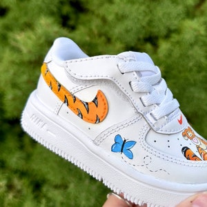 May include: White toddler's trainer with orange and black tiger stripe details on the Nike swoosh and heel. A blue butterfly and cartoon character are painted on the side. The shoe has white laces and a white sole.