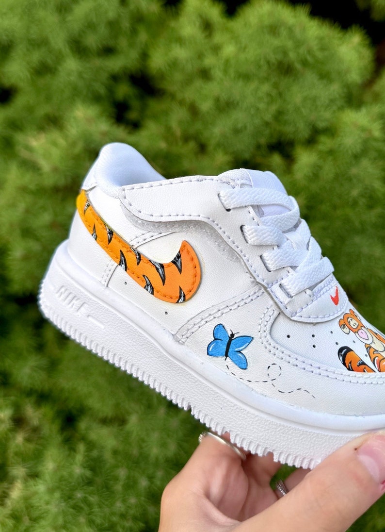 May include: White toddler's trainer with orange and black tiger stripe details on the Nike swoosh and heel. A blue butterfly and cartoon character are painted on the side. The shoe has white laces and a white sole.