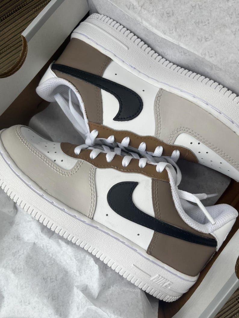 Puede incluir: Un par de zapatillas Air Force 1 personalizadas en una caja. Los zapatos presentan una base blanca con detalles en beige, marr&oacute;n y negro. El logo de Nike es negro. Las suelas y los cordones son blancos.