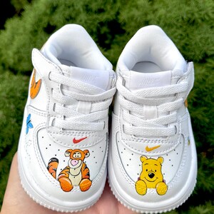 May include: White baby trainers decorated with cartoon characters Tigger and Winnie the Pooh. The shoes have a red Nike swoosh and a yellow swoosh. The trainers are decorated with orange, yellow, and blue accents.