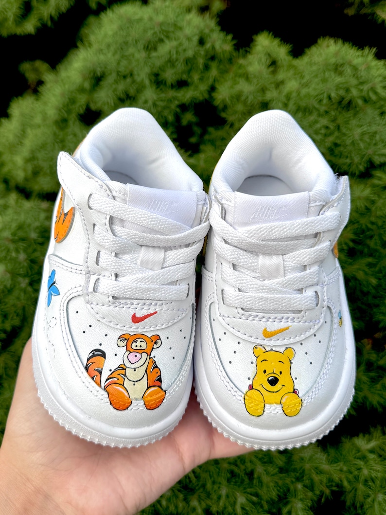 May include: White baby trainers decorated with cartoon characters Tigger and Winnie the Pooh. The shoes have a red Nike swoosh and a yellow swoosh. The trainers are decorated with orange, yellow, and blue accents.