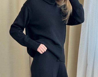 May include: A black long-sleeve sweater with a ribbed turtleneck collar and matching ribbed cuffs, paired with black wide-leg trousers. The sweater features a thick knit texture, while the trousers have a smooth, straight-leg silhouette.