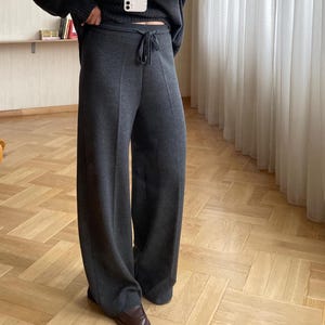 May include: A dark grey knit turtleneck sweater and matching wide-leg trousers. The trousers have a drawstring waist. The outfit is paired with brown leather shoes. The outfit is a casual, comfortable style.