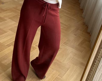 May include: A long-sleeved white crew neck sweater with a ribbed collar and cuffs paired with high-waisted, wide-leg knit trousers in a deep burgundy red color, featuring a drawstring waist and a smooth, solid-colored fabric texture.