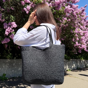 May include: A large, black woven tote bag with sturdy handles. The bag has a rectangular shape and a textured surface. The background features a vibrant display of purple flowers and a glimpse of a sunny day.