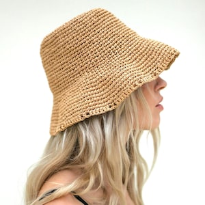 May include: A woven straw bucket hat in a natural beige color. The hat has a slightly downward-sloping brim and a textured weave. The hat is shown from a side view.