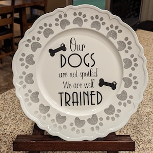 May include: White ceramic plate with a scalloped edge and a silver glitter paw print design. The plate has black text that reads "Our dogs are not spoiled. We are well trained."