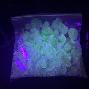 May include: A clear plastic bag filled with numerous, irregularly shaped, neon green crystalline solids. The bag is sealed with a zipper closure. The image is illuminated with a purple and green light, creating a glowing effect against a dark background.