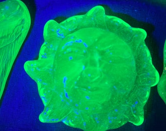 May include: A green glass sun with a face, with blue flecks throughout the glass. The sun is a decorative piece.