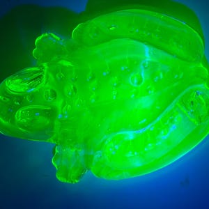 Puede incluir: Una figura de vidrio translúcido con forma de rana que brilla con una intensa luz verde neón. La superficie del vidrio presenta detalles moldeados, incluyendo protuberancias en relieve a lo largo del lomo y extremidades definidas, capturando un efecto luminoso.