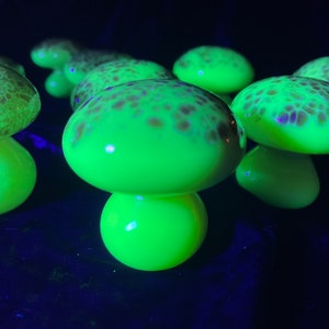 May include: A collection of glowing glass mushroom sculptures. The mushrooms have a bright neon green color with brown spotted caps. The stems are rounded and the caps are domed. The background is dark.