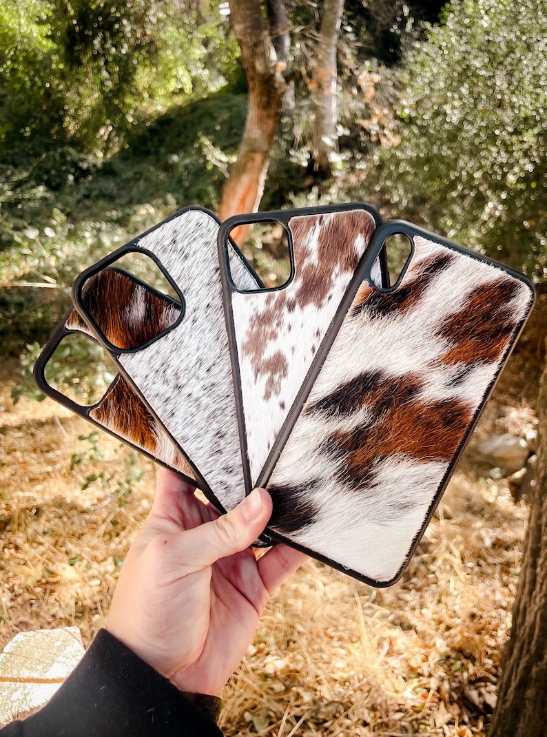 May include: Four different phone cases with a cowhide pattern. The cases are black with a white and brown cowhide pattern. The cases are arranged in a fan shape.