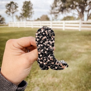 May include: A black and gold glitter cowboy boot charm. The boot is made of a sparkly material and has a pointed toe.