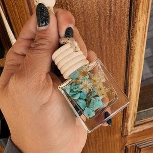 Puede incluir: Una botella de vidrio transparente de forma cuadrada llena de piedras turquesa y flores blancas secas. La botella está unida a una cuenta de madera blanca y una cuerda de hilo.