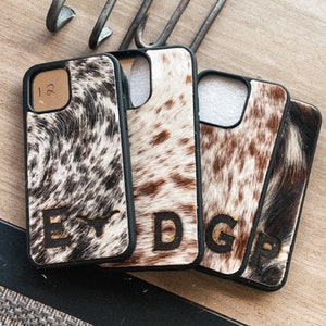 May include: Four black phone cases with a cowhide pattern and a letter initial on each. The cases are arranged in a row on a wooden surface.