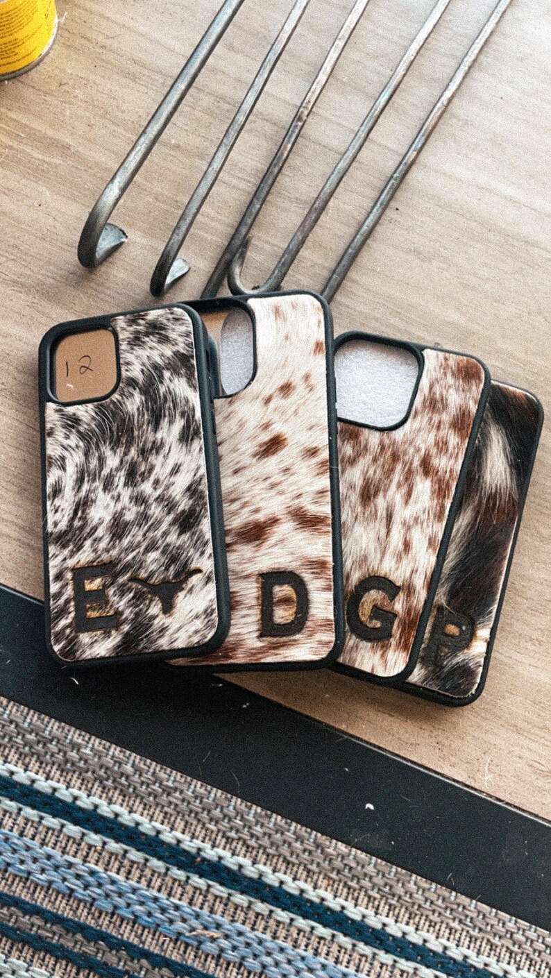 May include: Four black phone cases with a cowhide pattern and a letter initial on each. The cases are arranged in a row on a wooden surface.