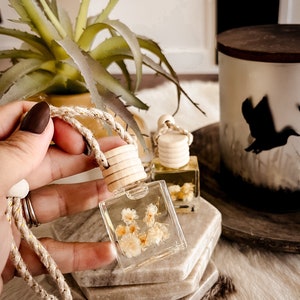 Puede incluir: Dos pequeñas botellas de vidrio con tapas de corcho y flores blancas en su interior. Las botellas cuelgan de una cuerda y están sobre una pila de posavasos beige.