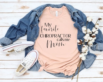 以下が含まれることがあります： 桃色のTシャツに、黒い筆記体で「My favorite chiropractor calls me Mom」という文字が書かれています。