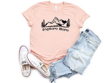 以下が含まれることがあります： ピーチ色のTシャツに、黒い山脈のグラフィックと「Explore More」の文字がプリントされています。このTシャツは、ライトブルーのデニムジーンズと白いスニーカーと合わせています。